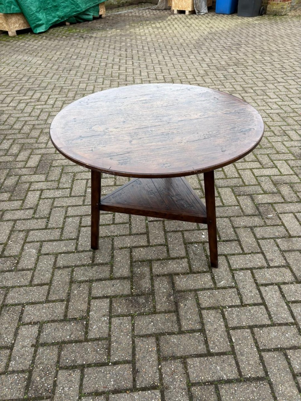 An antique dark oak cricket table featuring a circular plank top, three splayed legs, and a lower triangular shelf.