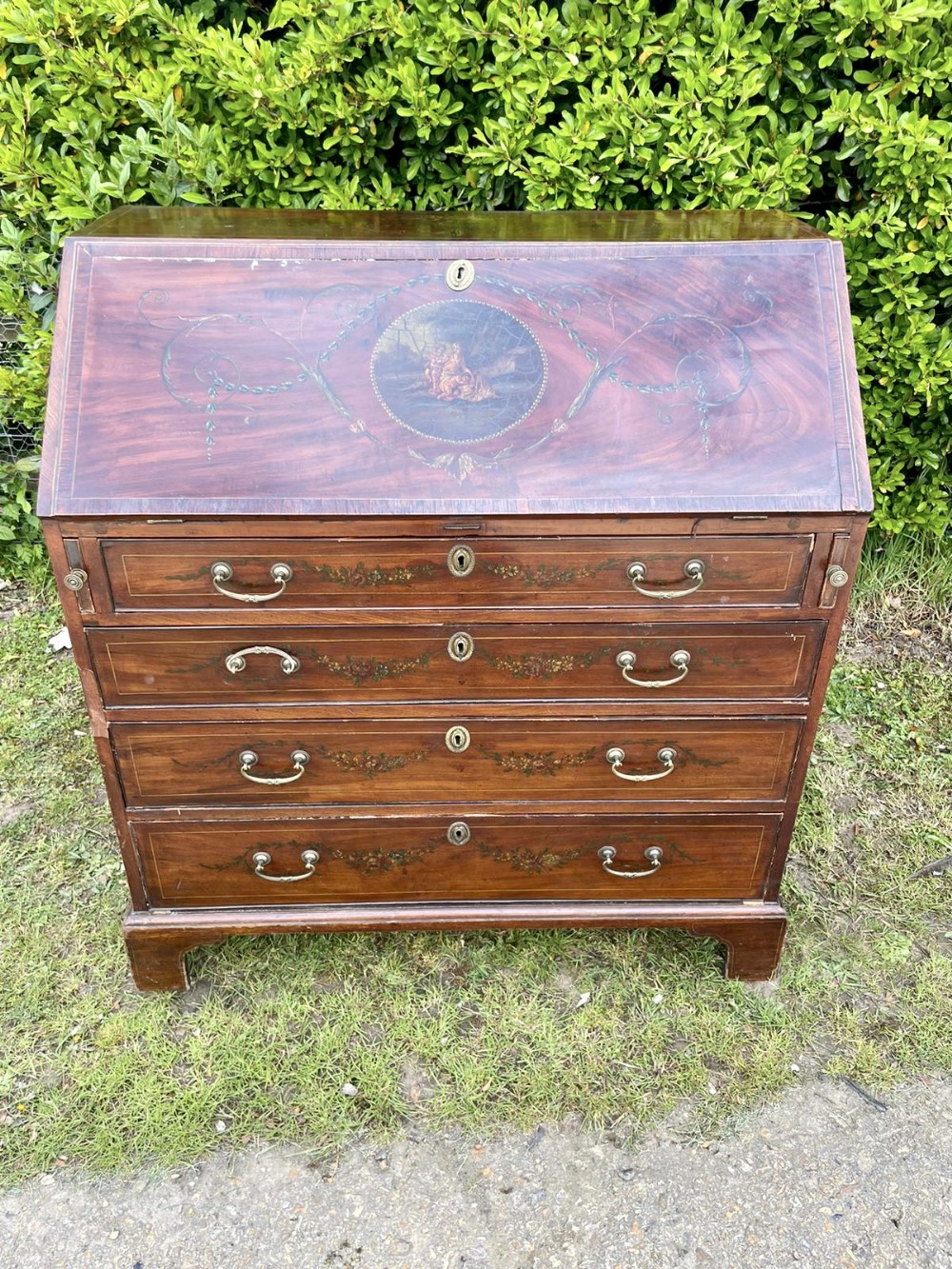 An antique George III mahogany slant-front bureau featuring hand-painted classical medallions and floral swags across four graduated drawers with brass swan-neck handles.