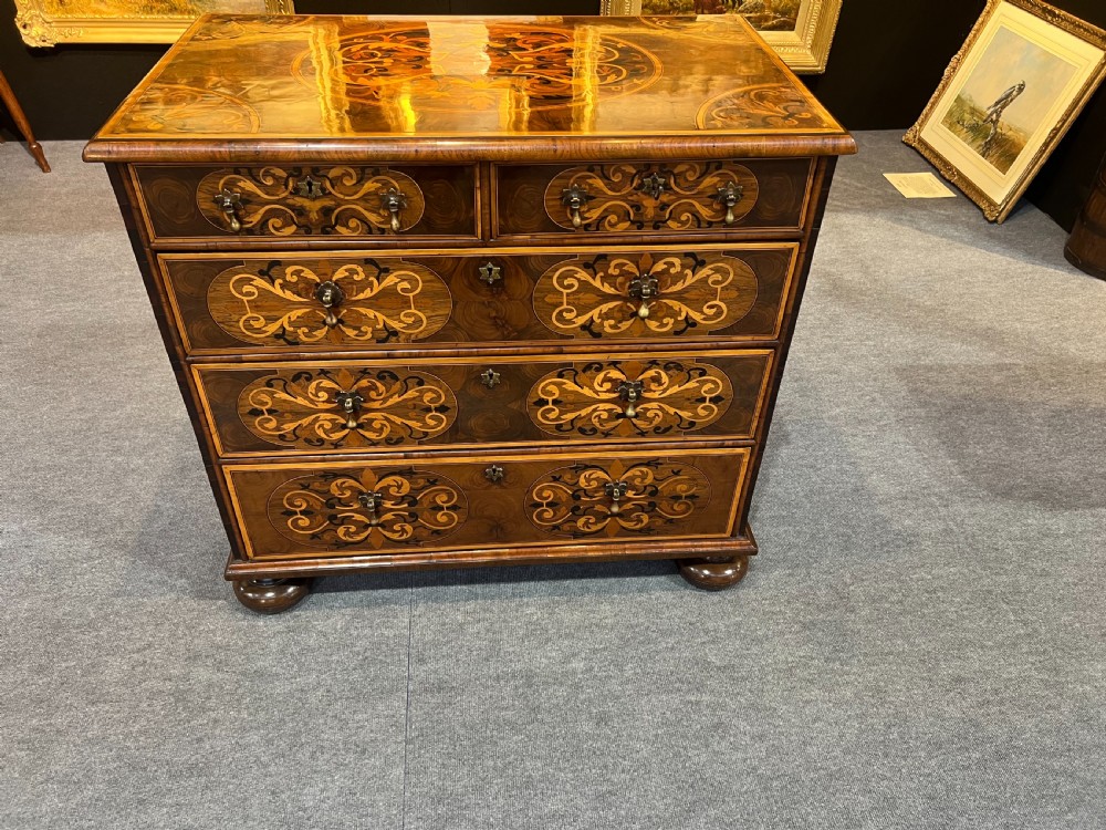 A walnut chest of drawers featuring intricate floral marquetry inlay across the drawer fronts and top surface, fitted with brass drop handles and raised on bun feet.