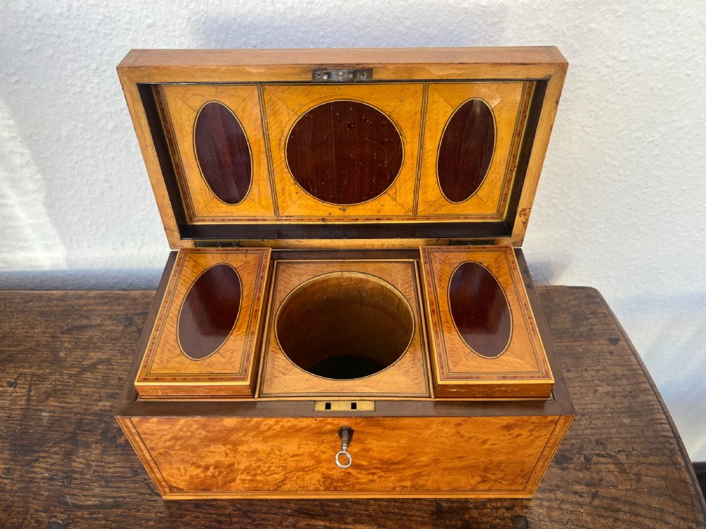 An open antique golden satinwood tea caddy displaying decorative marquetry ovals, two fitted side compartments, a central mixing aperture, and a key.