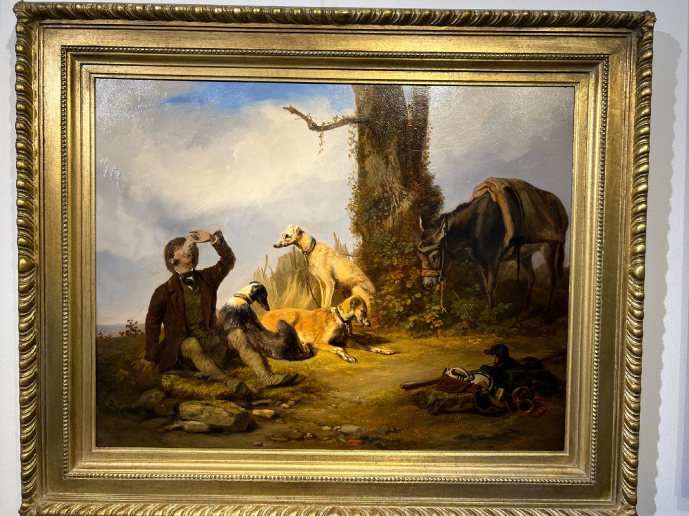 A nineteenth-century oil painting in a carved gold frame depicting a seated huntsman drinking alongside three resting hounds and a saddled donkey.