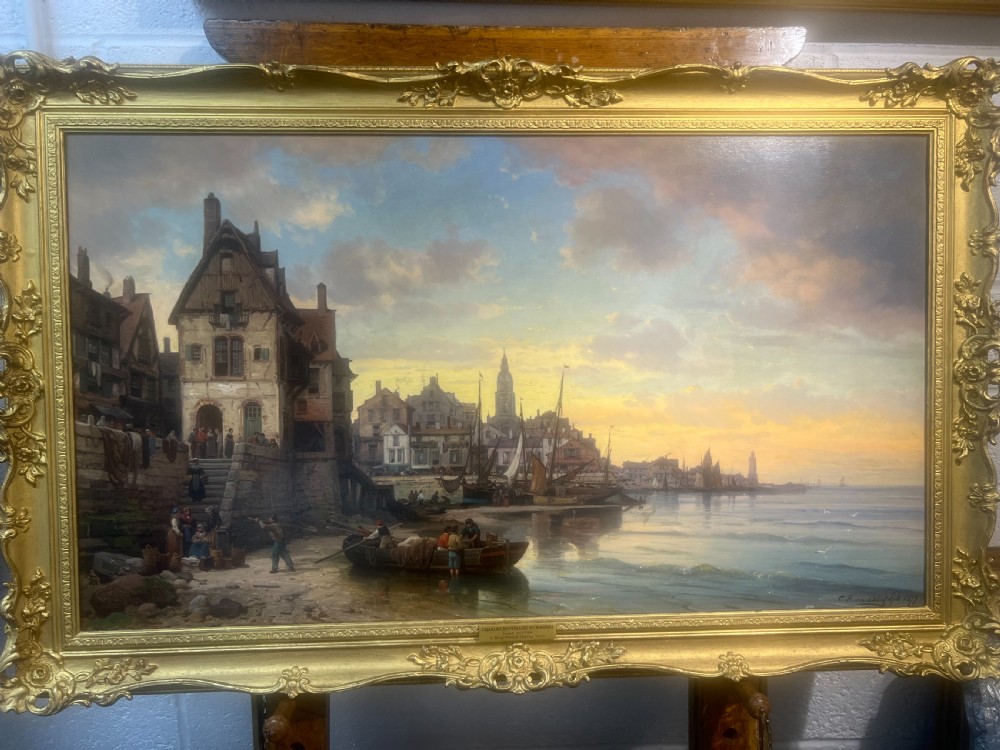 A 19th-century French oil painting depicting a coastal harbour town at sunset, presented within a heavily carved giltwood frame.