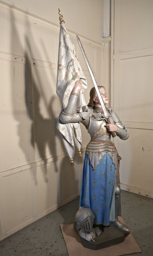 fig 1. A French polychrome plaster statue depicting Saint Joan of Arc in silver armour and a blue tunic decorated with fleur-de-lis, holding a sword and flag.