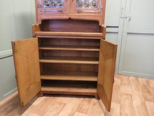 fig 1. A solid golden oak Arts and Crafts dresser featuring upper bullseye leaded glass doors with iron strap hinges above an open shelf and carved panelled lower doors.