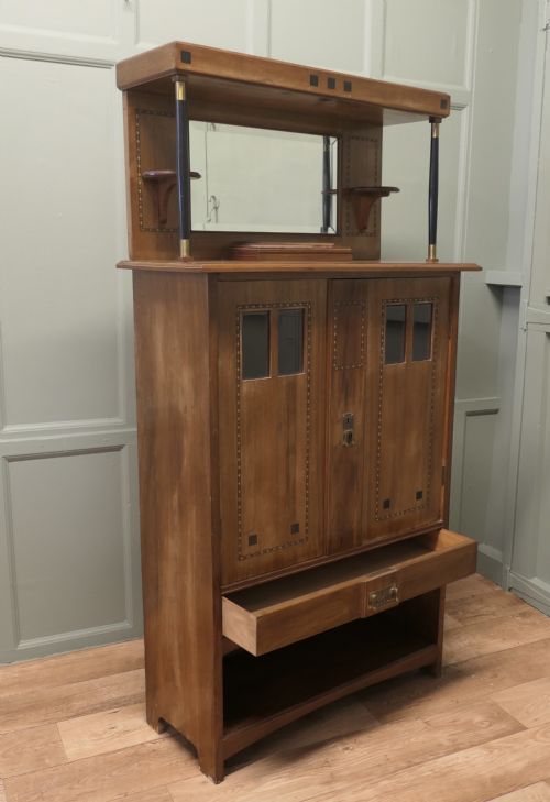 fig 1. A Secessionist warm brown oak cocktail cabinet featuring a mirror-backed upper shelf supported by dark pillars, geometric wood inlay detailing on double doors, brass keyhole fittings, and an open lower shelf gallery.