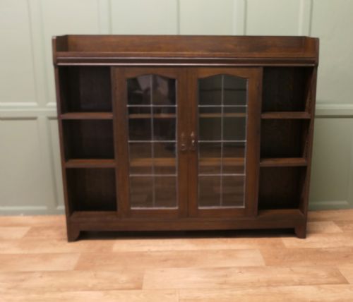 fig 1. An Arts and Crafts dark oak bookcase featuring central leaded glass double doors flanked by open tier side shelves and a raised top gallery.