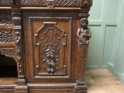 fig 1. A heavily carved warm brown oak sideboard with three top drawers, relief carved cupboard doors, putti side supports, and a central open recess.