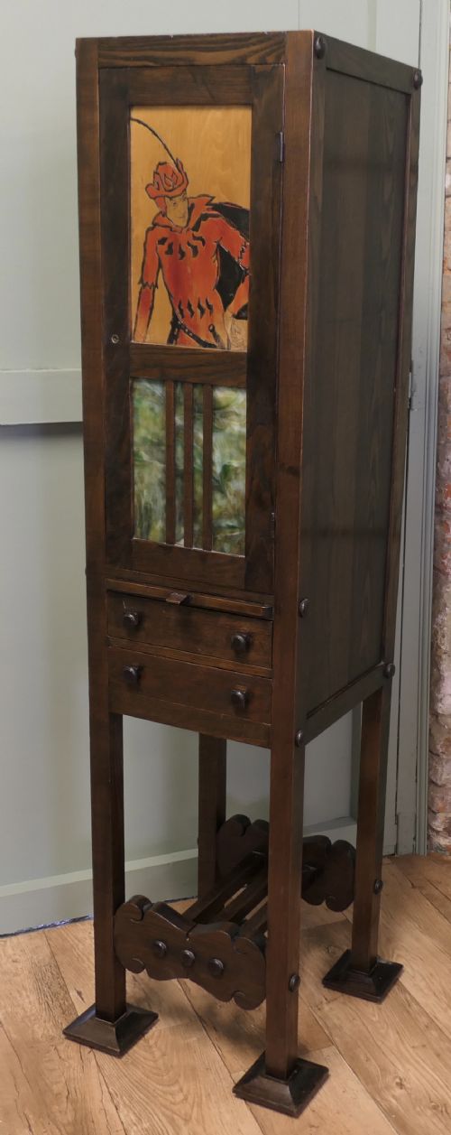fig 1. A tall, slender dark oak Arts and Crafts cabinet featuring a decorative inlaid wood panel depicting a figure in red, a slatted glass door section, two small drawers, and a lower shelf.