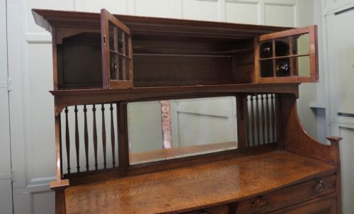 fig 1. A large Arts and Crafts oak sideboard featuring a mirrored back, upper leaded glass cabinets, carved lower cupboard doors with botanical motifs, and brass handles.