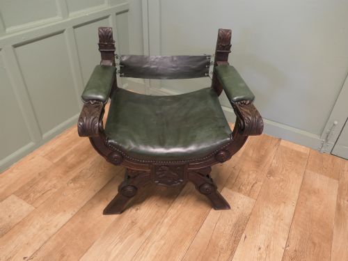 fig 1. A 19th-century carved oak X-frame armchair featuring green leather upholstery, acanthus finials, and a front crossbar carved with a lion shield.