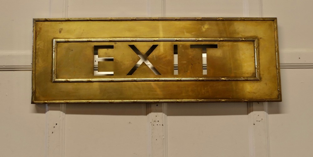 A large rectangular brass wall sign featuring stencil cut-out lettering spelling EXIT framed by textured borders.