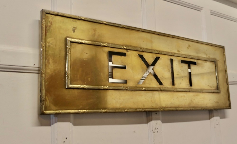 A large rectangular brass wall sign featuring stencil cut-out lettering spelling EXIT framed by textured borders. View II