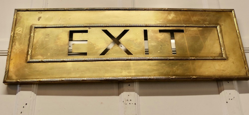 A large rectangular brass wall sign featuring stencil cut-out lettering spelling EXIT framed by textured borders. View IV