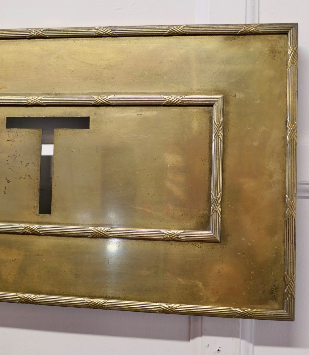 A large rectangular brass wall sign featuring stencil cut-out lettering spelling EXIT framed by textured borders. View V