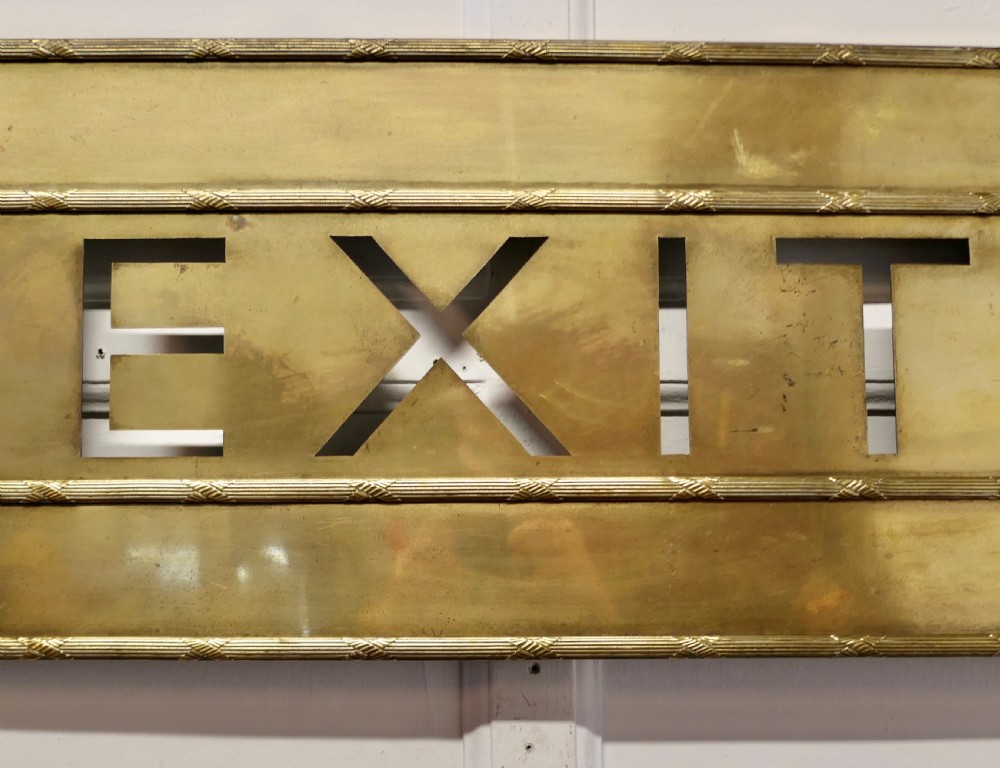 A large rectangular brass wall sign featuring stencil cut-out lettering spelling EXIT framed by textured borders. View VI