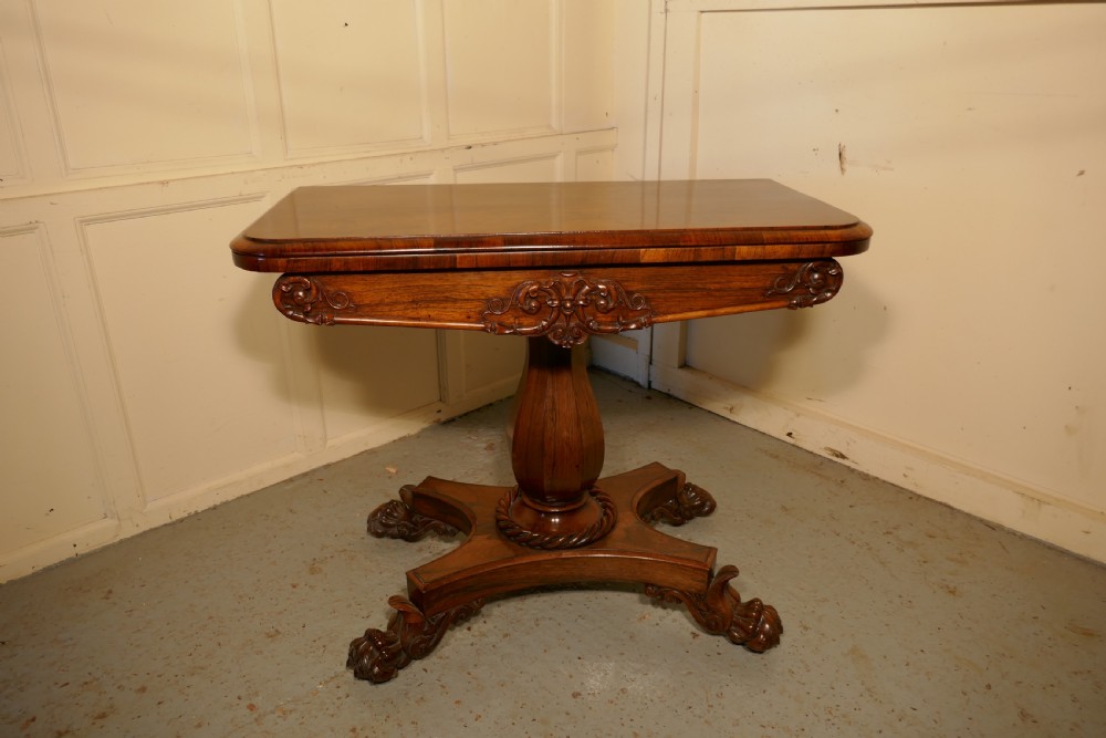 A William IV dark wood folding card table featuring a carved frieze, central turned pedestal, and a concave quadform plinth base with scroll feet.