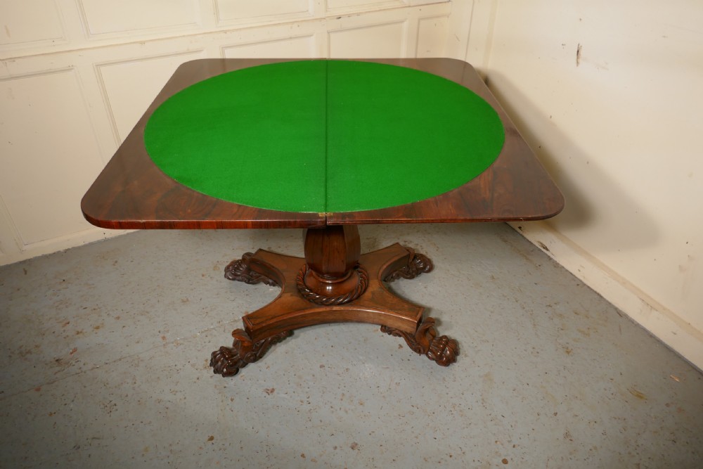 A William IV dark wood folding card table featuring a carved frieze, central turned pedestal, and a concave quadform plinth base with scroll feet. View X