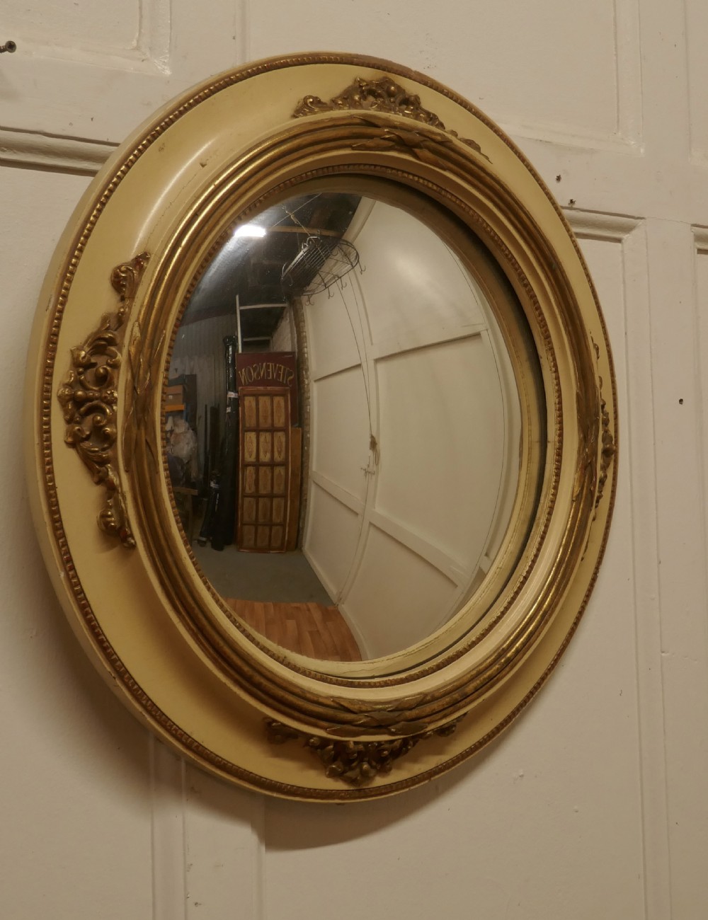 An oval French convex wall mirror featuring a painted cream frame accented with gilded acanthus leaf embellishments.