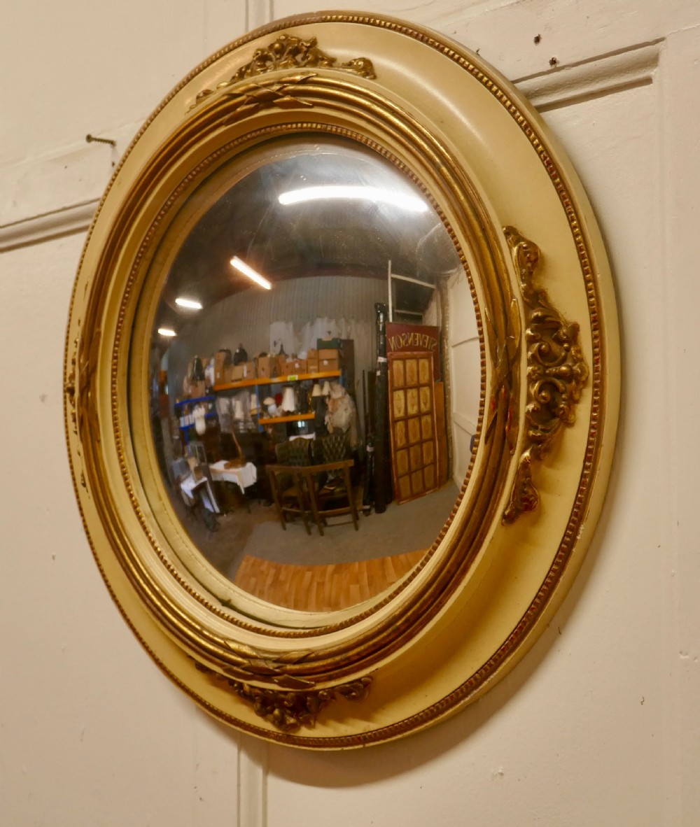 An oval French convex wall mirror featuring a painted cream frame accented with gilded acanthus leaf embellishments. View IV