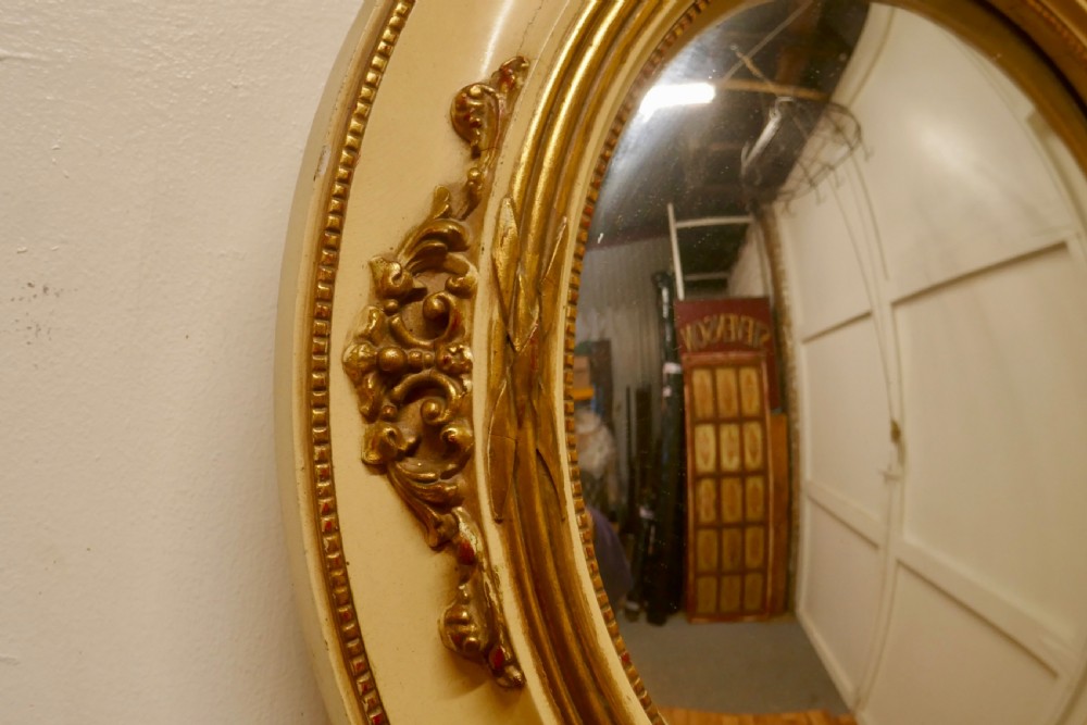 An oval French convex wall mirror featuring a painted cream frame accented with gilded acanthus leaf embellishments. View VI