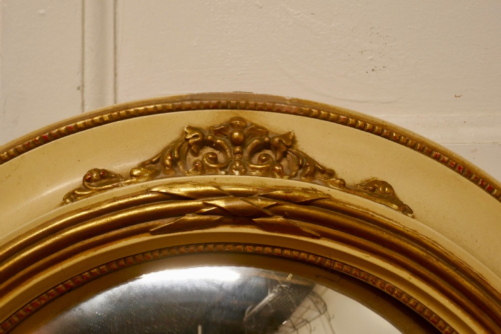 An oval French convex wall mirror featuring a painted cream frame accented with gilded acanthus leaf embellishments. View VII