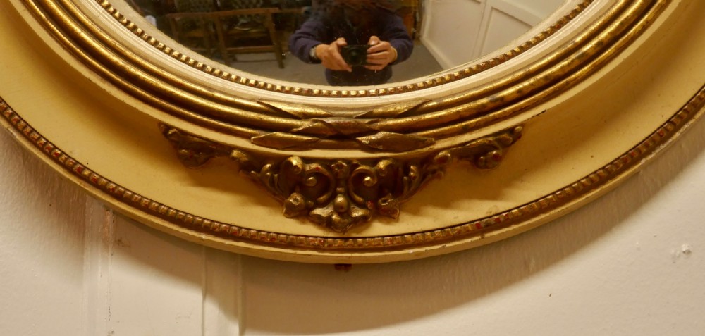 An oval French convex wall mirror featuring a painted cream frame accented with gilded acanthus leaf embellishments. View VIII