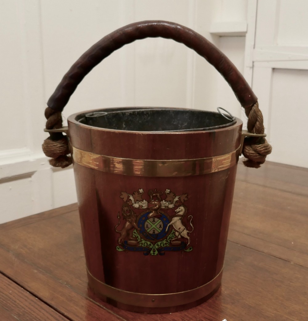 A 19th-century dark oak fire bucket featuring brass binding, a thick rope handle covered in brown leather, and a painted Plymouth armorial crest on the front.