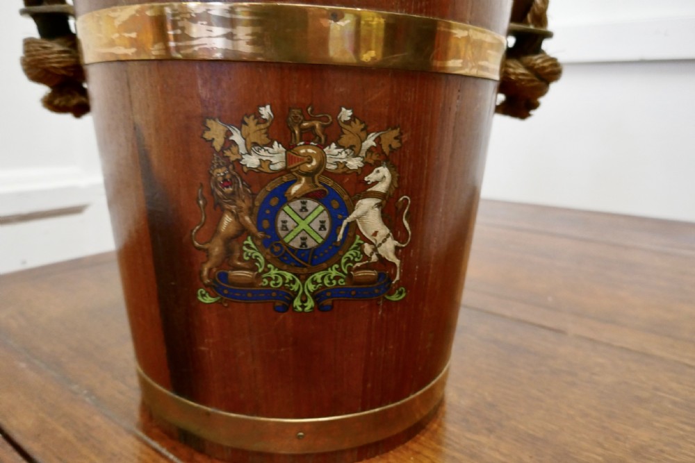 A 19th-century dark oak fire bucket featuring brass binding, a thick rope handle covered in brown leather, and a painted Plymouth armorial crest on the front. View II