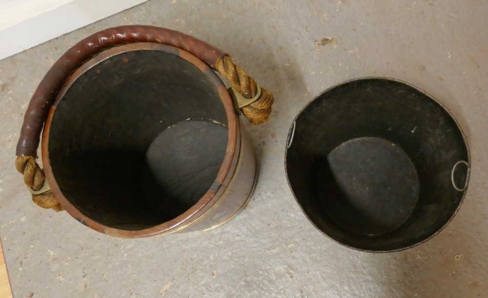 A 19th-century dark oak fire bucket featuring brass binding, a thick rope handle covered in brown leather, and a painted Plymouth armorial crest on the front. View VI