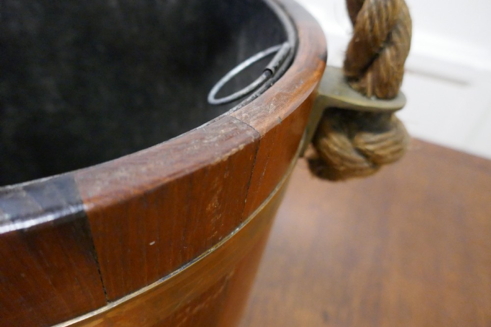 A 19th-century dark oak fire bucket featuring brass binding, a thick rope handle covered in brown leather, and a painted Plymouth armorial crest on the front. View VIII
