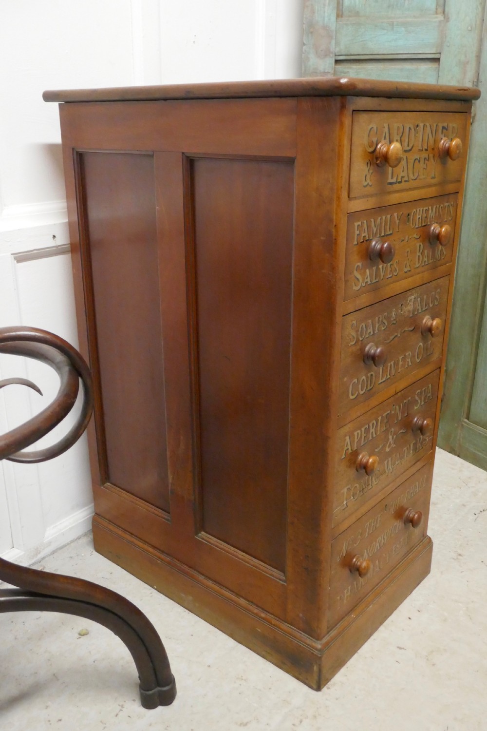 A tall Victorian pine chest featuring five drawers with turned wooden knobs and hand-painted gilt pharmacy lettering across each drawer front. View XII