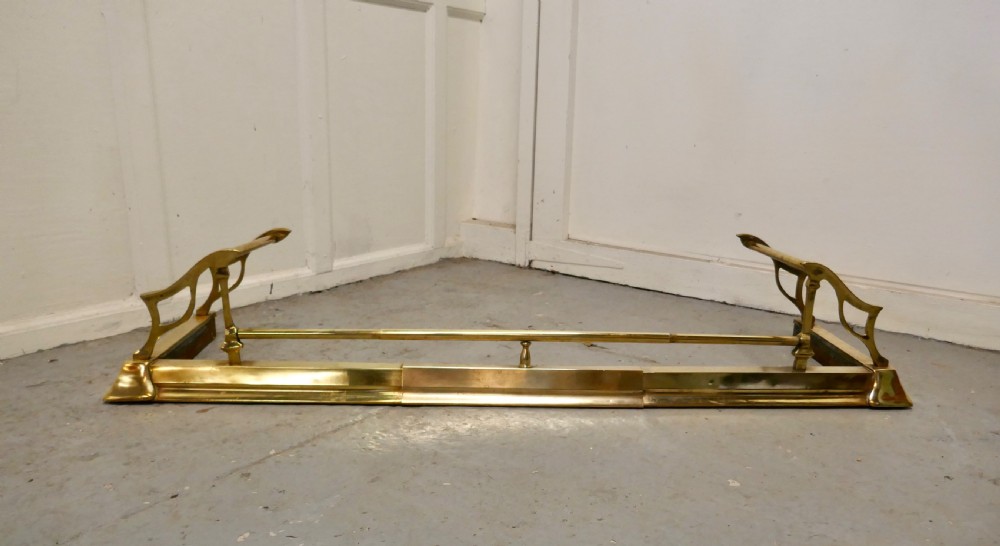 A polished Art Nouveau brass fireplace fender featuring sinuous openwork side supports and a central low horizontal rail.