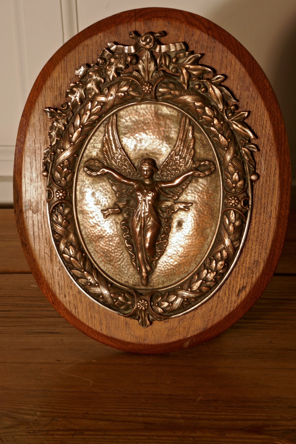 An oval oak wooden plaque featuring a copper relief medallion depicting the winged goddess Nike surrounded by a laurel wreath.