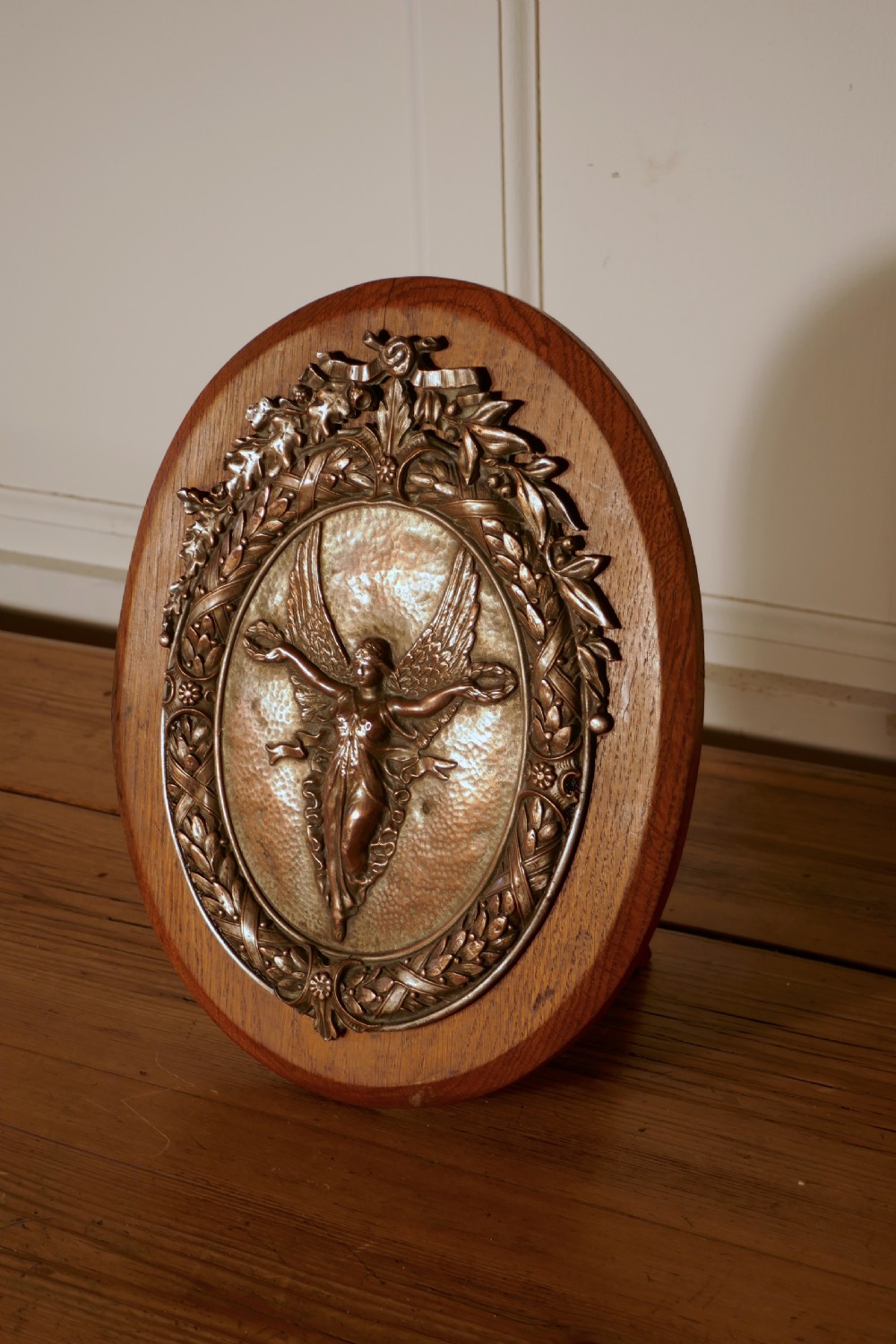 An oval oak wooden plaque featuring a copper relief medallion depicting the winged goddess Nike surrounded by a laurel wreath. View III