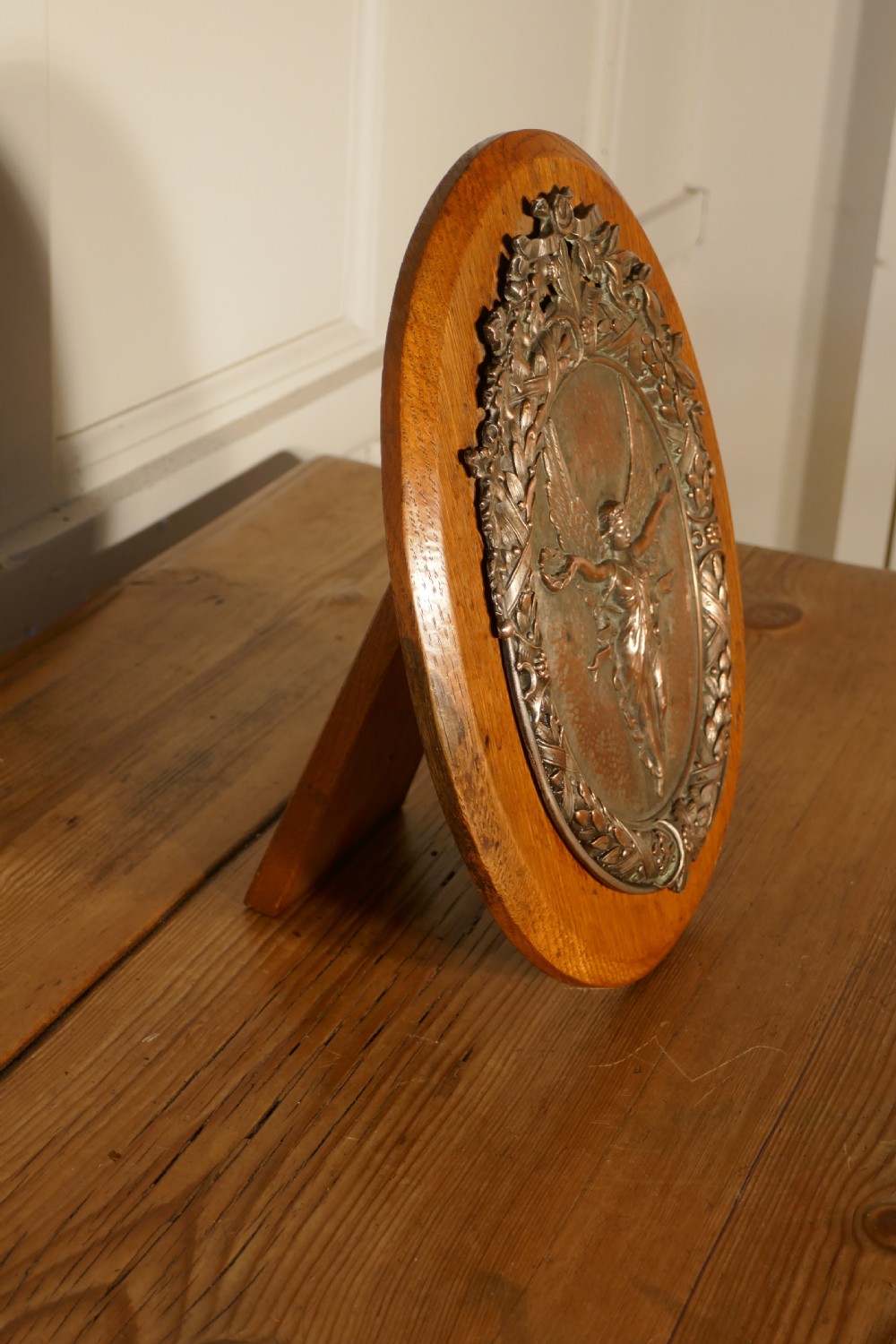 An oval oak wooden plaque featuring a copper relief medallion depicting the winged goddess Nike surrounded by a laurel wreath. View V