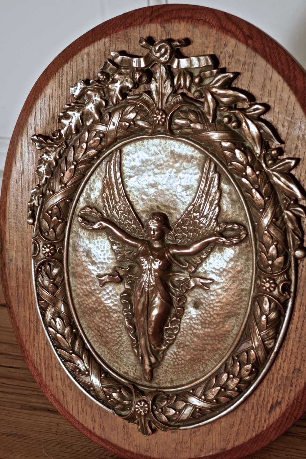 An oval oak wooden plaque featuring a copper relief medallion depicting the winged goddess Nike surrounded by a laurel wreath. View VI