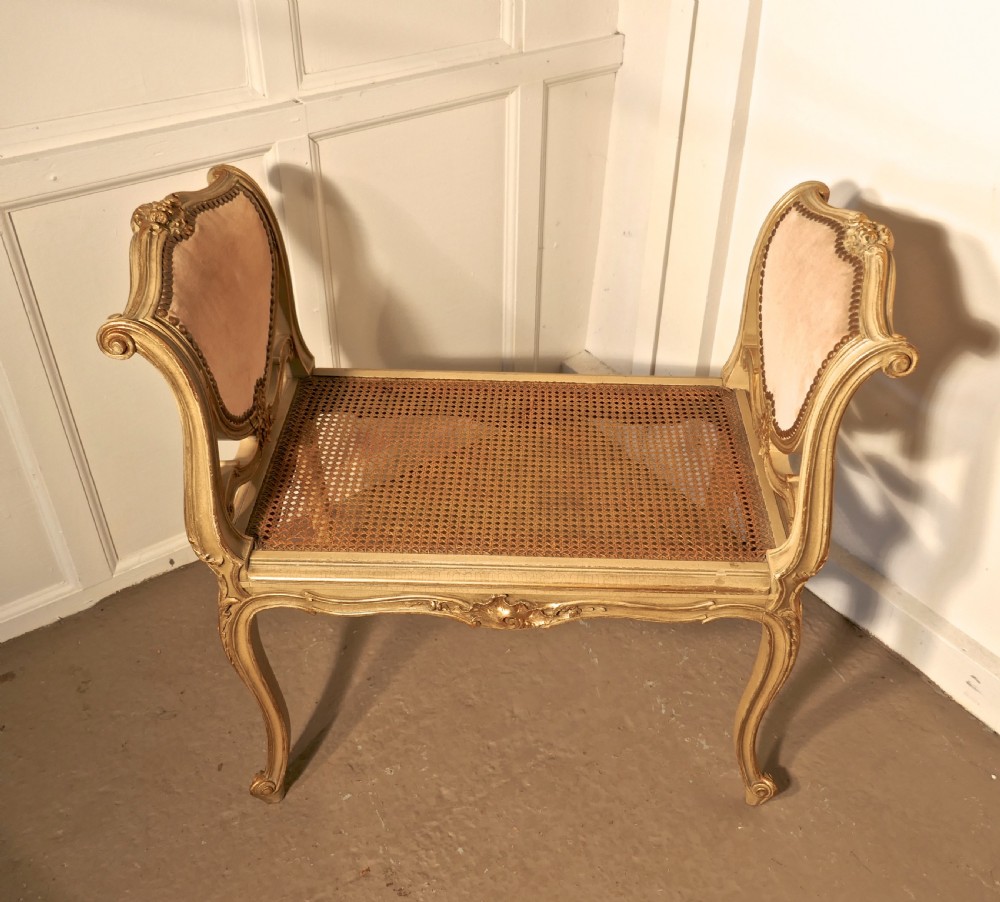 An antique French giltwood window seat featuring raised dusky pink velvet ends with studding, a cane seat, and curved cabriole legs.