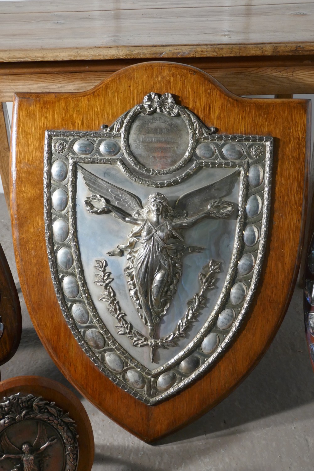 A shield-shaped oak wall plaque featuring a silver-plated relief of the winged goddess Nike surrounded by laurel motifs and small engraved oval shields.