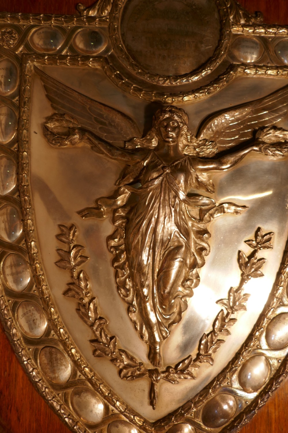 A shield-shaped oak wall plaque featuring a silver-plated relief of the winged goddess Nike surrounded by laurel motifs and small engraved oval shields. View IV