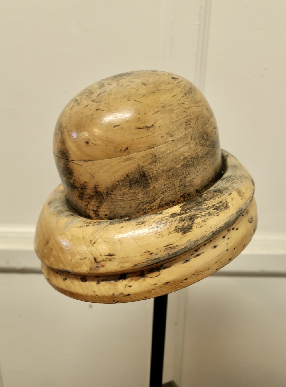 A carved light fruitwood hat block featuring a rounded crown and brim section, mounted on a dark metal stem with a square dark wood base. View II