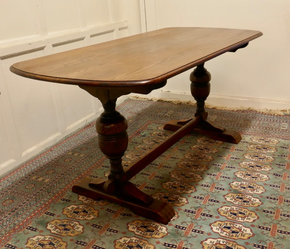 A solid warm brown oak refectory dining table featuring turned baluster supports connected by a low central stretcher base. View IX