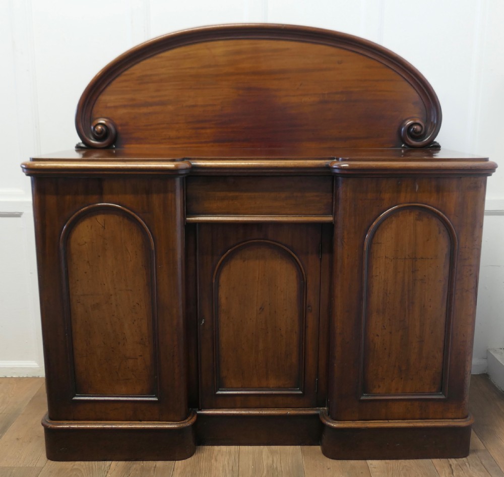A Victorian mahogany sideboard featuring a large arched scroll backboard over a recessed central cupboard door flanked by panelled side cupboards on a plinth base.