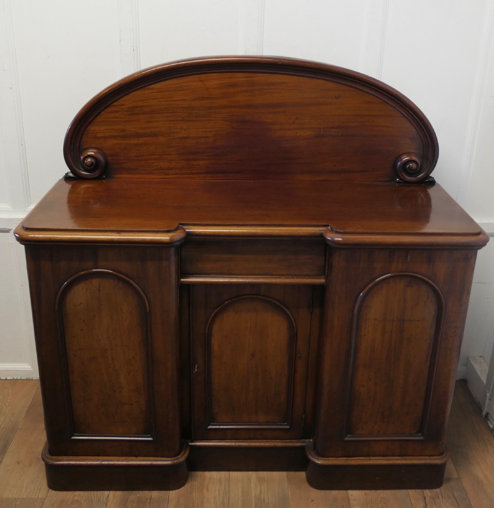 A Victorian mahogany sideboard featuring a large arched scroll backboard over a recessed central cupboard door flanked by panelled side cupboards on a plinth base. View V