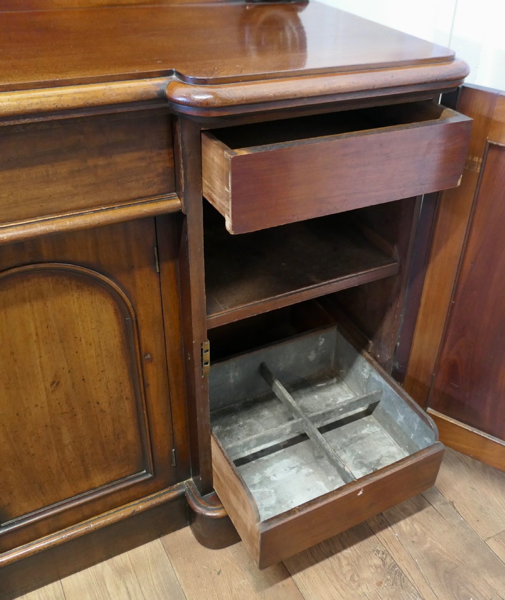 A Victorian mahogany sideboard featuring a large arched scroll backboard over a recessed central cupboard door flanked by panelled side cupboards on a plinth base. View VI