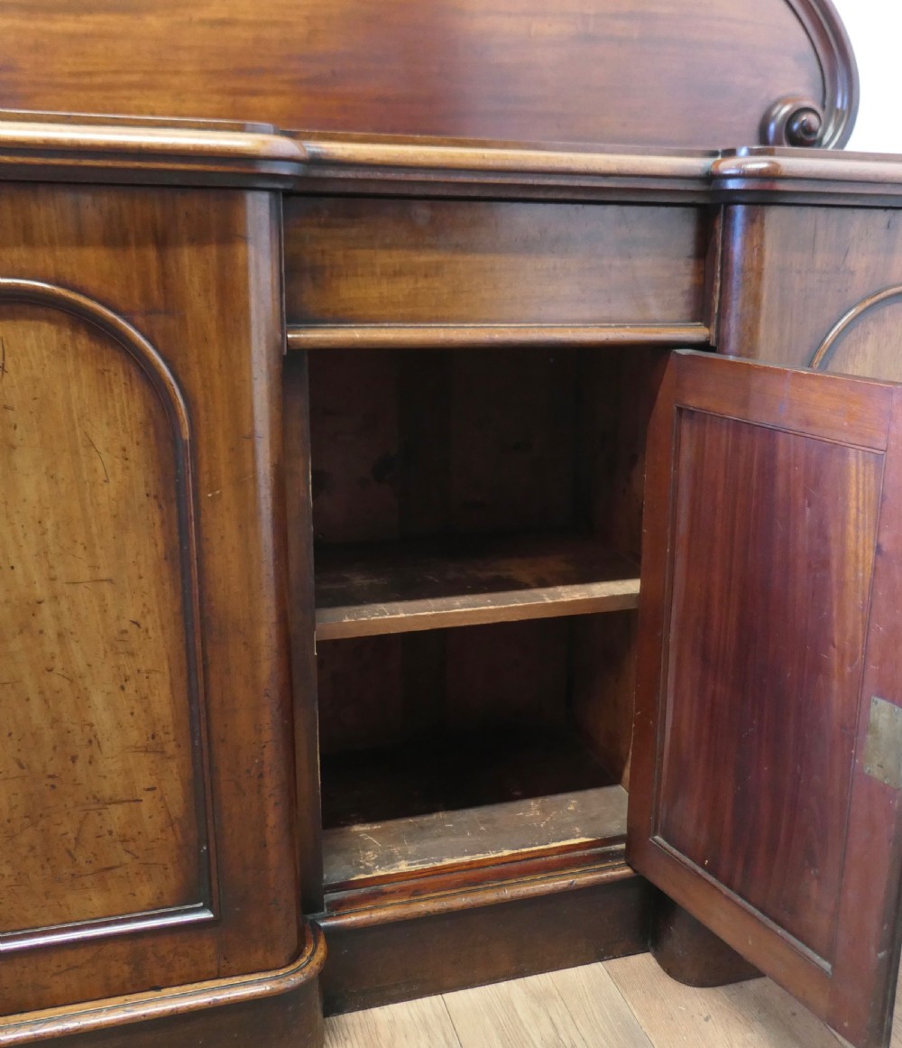 A Victorian mahogany sideboard featuring a large arched scroll backboard over a recessed central cupboard door flanked by panelled side cupboards on a plinth base. View IX
