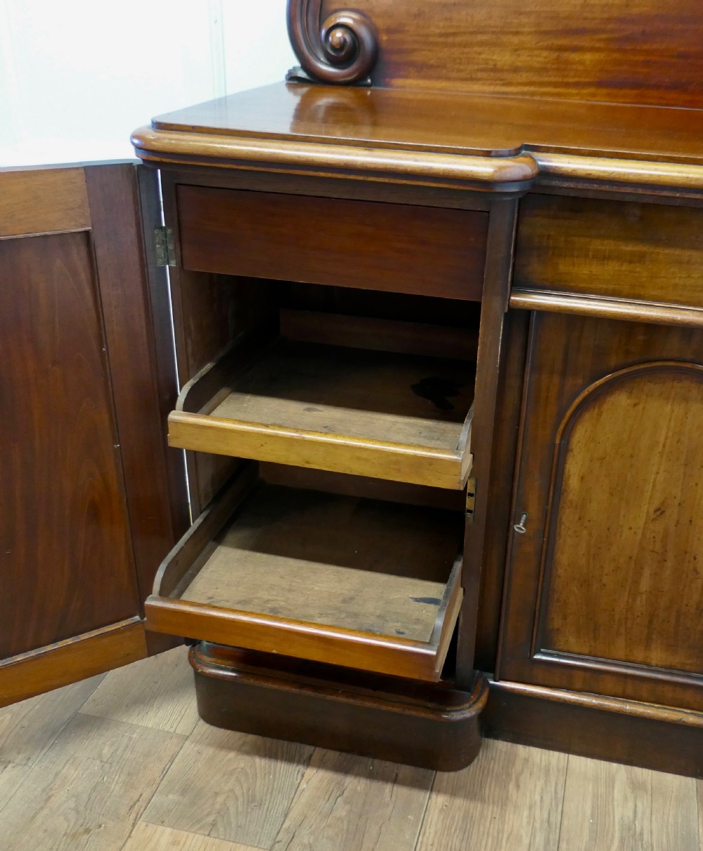 A Victorian mahogany sideboard featuring a large arched scroll backboard over a recessed central cupboard door flanked by panelled side cupboards on a plinth base. View X