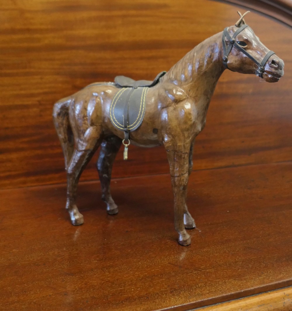 A handcrafted brown leather model of a horse featuring a miniature dark leather saddle, harness, and brass stirrup loops.