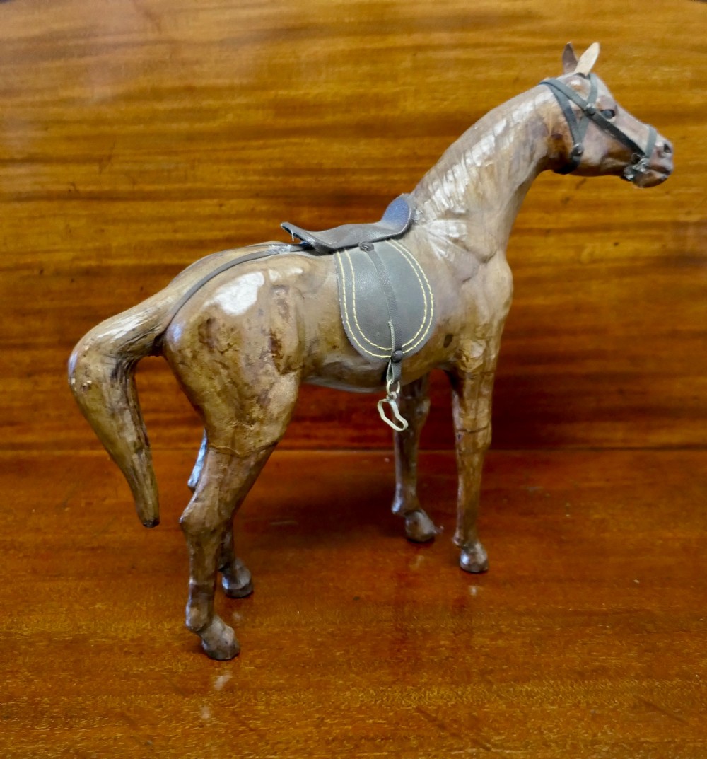 A handcrafted brown leather model of a horse featuring a miniature dark leather saddle, harness, and brass stirrup loops. View II