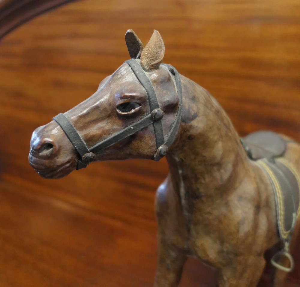 A handcrafted brown leather model of a horse featuring a miniature dark leather saddle, harness, and brass stirrup loops. View V