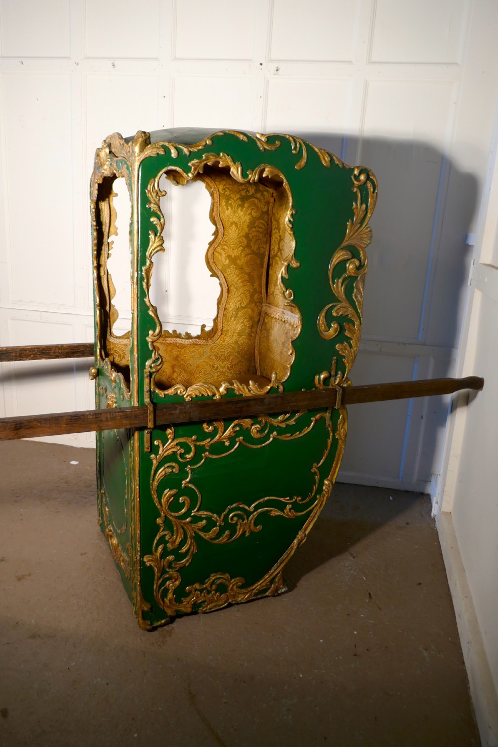 A 19th-century green painted and giltwood Italian sedan chair featuring hand-carved scrollwork, window openings, gold fabric lining, and wooden carrying poles.